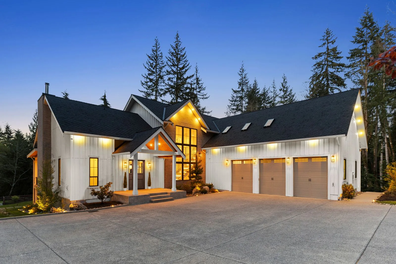 White gabled home at twilight with warm windows, a three-car garage and tall trees